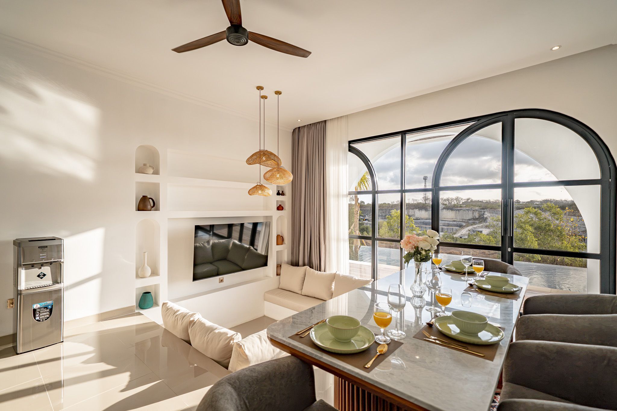 Una sala moderna con televisor de pantalla plana, zona de comedor con mesa de vidrio y un gran ventanal con vistas al paisaje.