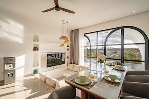 A modern living room with a flat-screen TV, a dining area with a glass table, and a large arched window offering a view of the landscape. - Jing Jang Villa Ungasan (Kutuh)