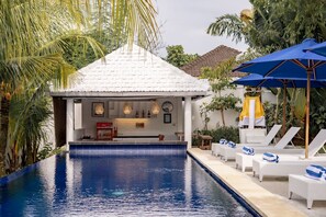 A pool area with lounge chairs and umbrellas, a bar area with a white tiled roof, and tropical plants. - Jing Jang Villa Ungasan (Kutuh)