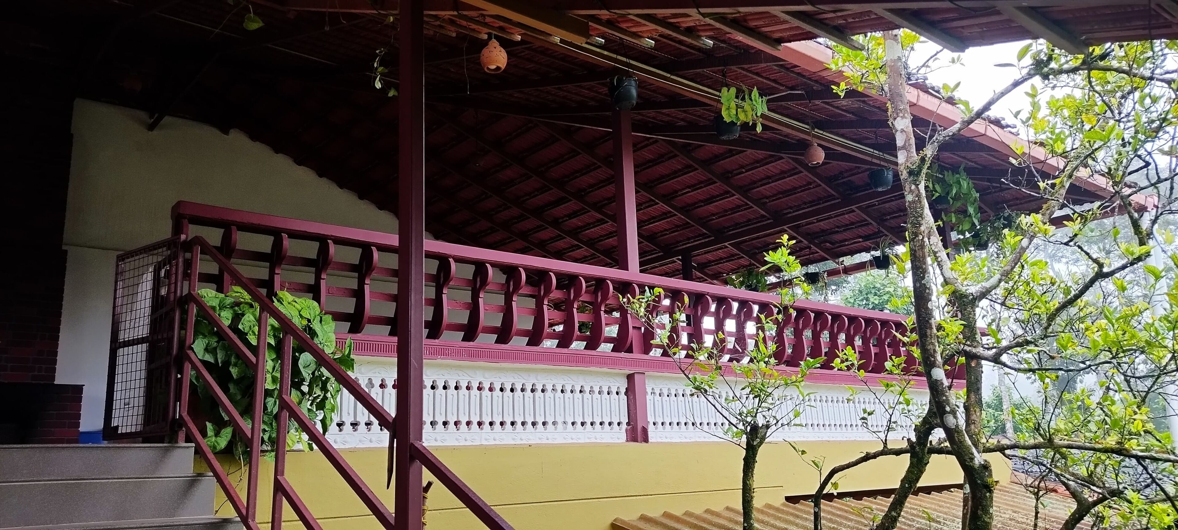 Un espacio al aire libre cubierto con una escalera de madera que lleva a un balcón, rodeado de vegetación y un edificio con fachada blanca y roja.
