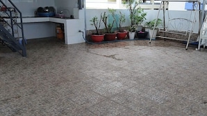 Ένα πλακόστρωτο αίθριο με γλάστρες γεμάτες φυτά, μια μεταλλική κρεμάστρα-καρέκλα και ένα λευκό παγκάκι.