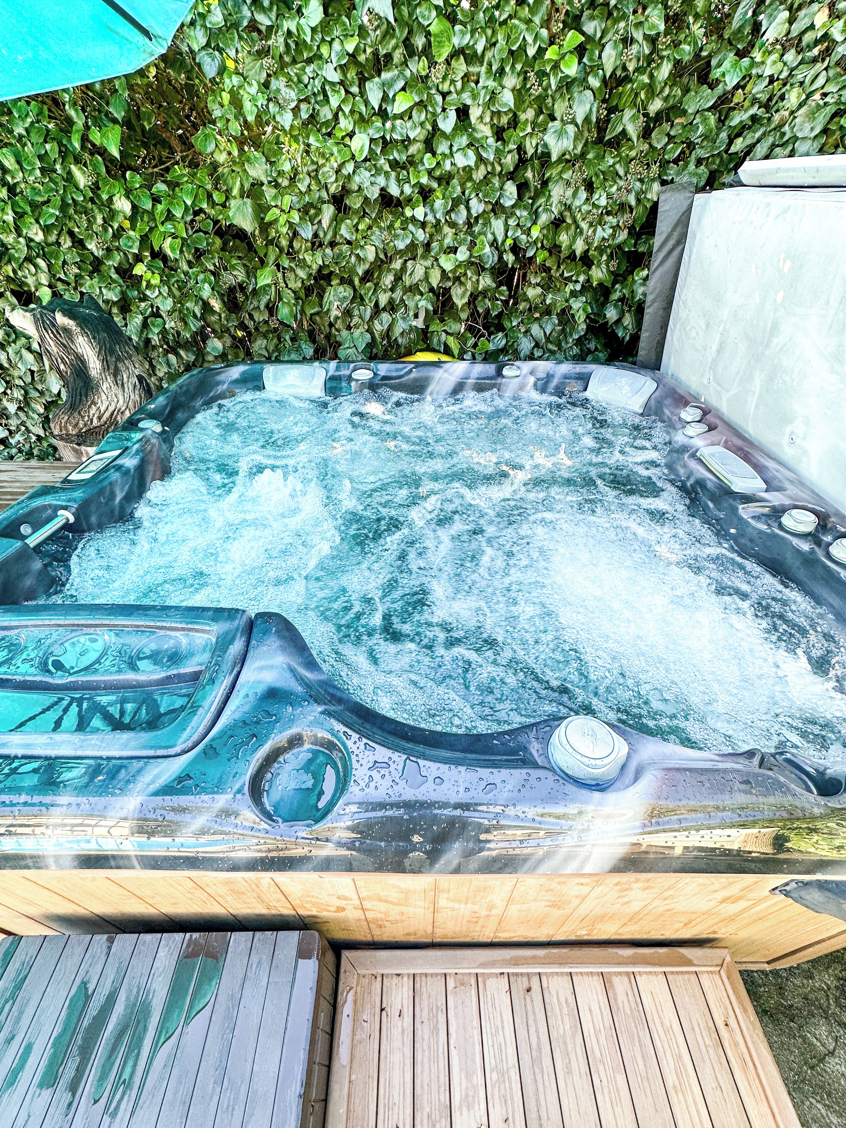 A hot tub with multiple jets, surrounded by a wooden deck and a hedge.