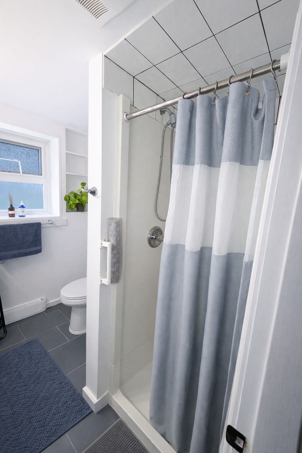 A shower with a grey and white striped shower curtain, a white toilet, a window with a view, and a blue rug.