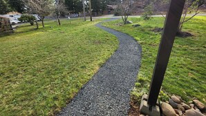 A gravel path curving through a grassy area with trees and a building in the background. - Comfortable Home in the Comox Valley (Comox)