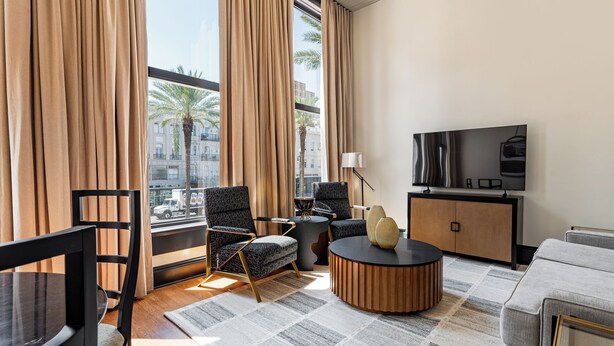 Habitación de hotel moderna con un ventanal grande, un televisor de pantalla plana, una mesa de centro redonda, un sofá y un comedor con una silla y una mesa.