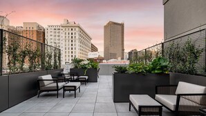 Une terrasse sur le toit, aménagée avec des espaces de détente en plein air, des plantes en pot et offrant une vue sur la ville au coucher du soleil.