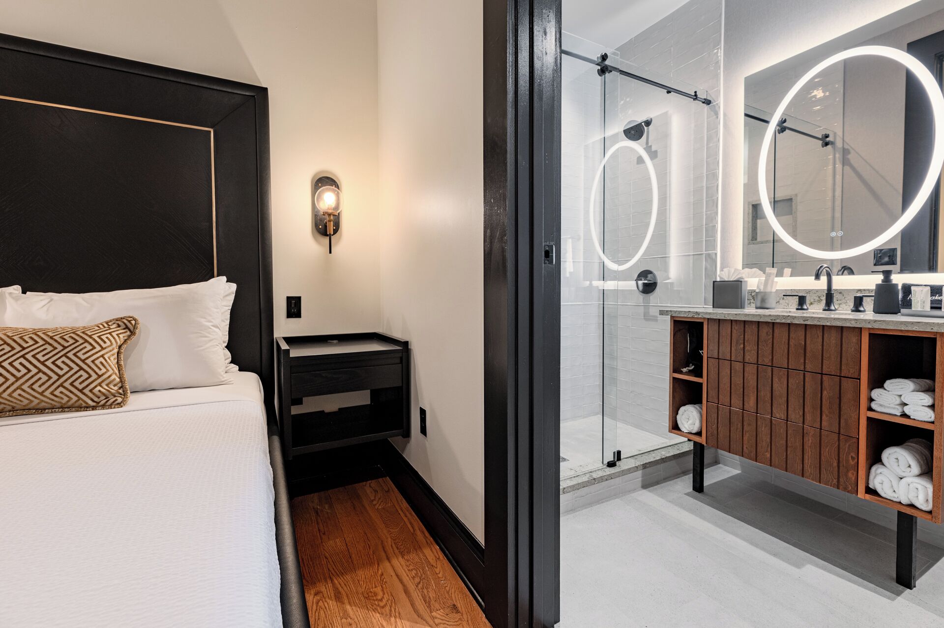 A modern hotel room with a bed, a nightstand, a bathroom with a large mirror, and a wooden vanity.