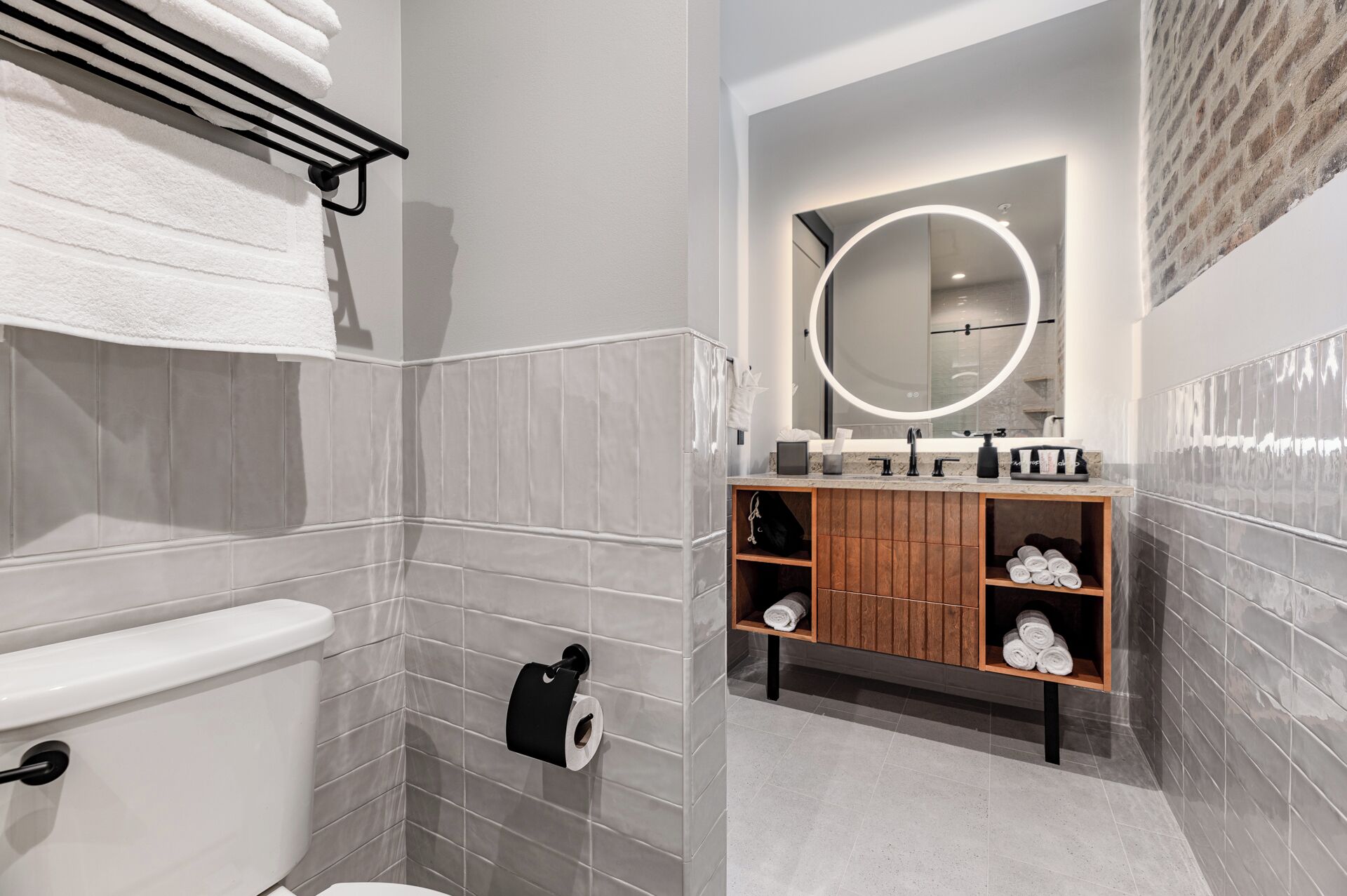 A modern bathroom with grey tiled walls, a toilet, a sink with a mirror, and shelves with towels.