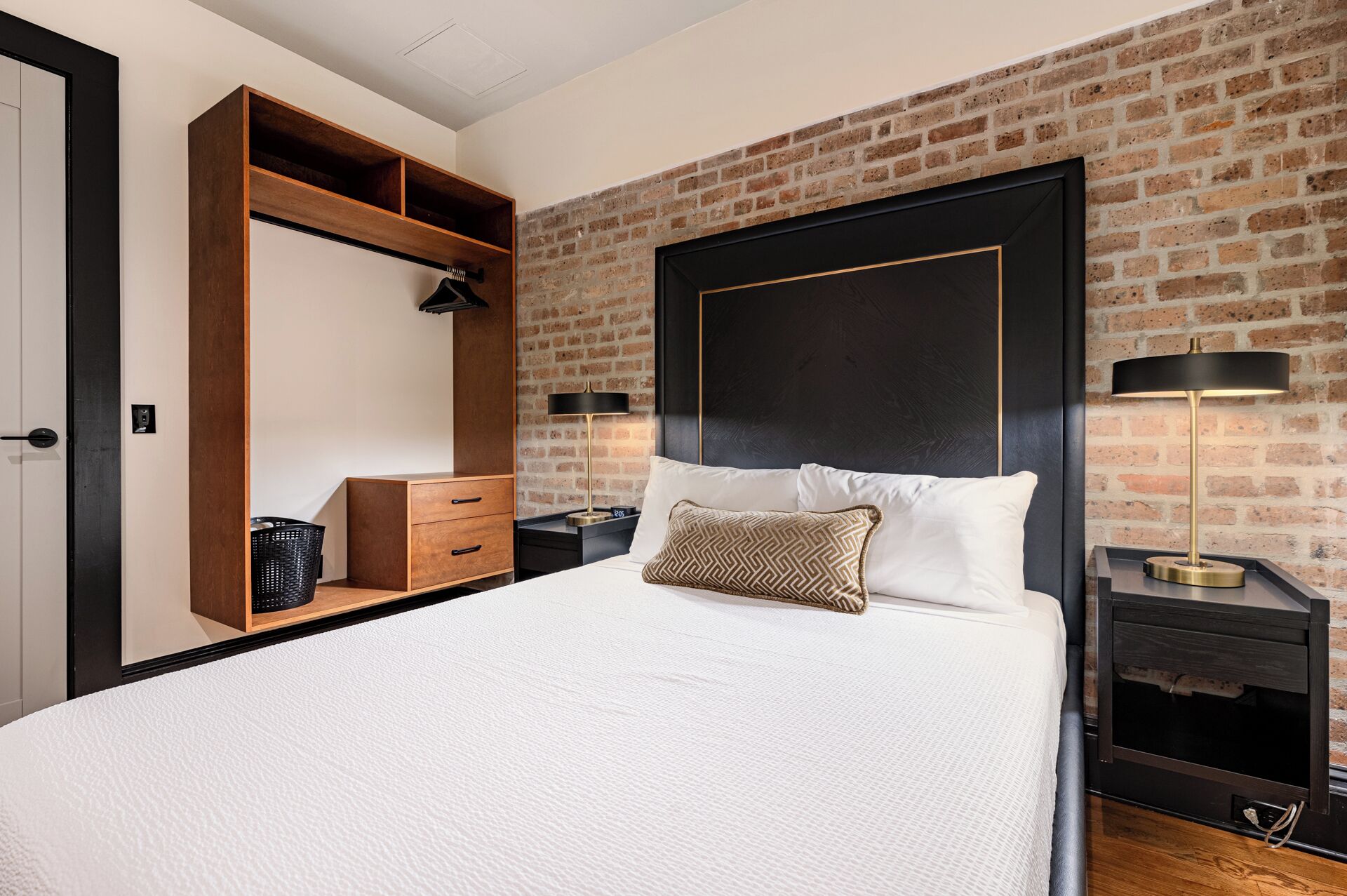 A modern bedroom with a brick wall, a large bed, a wooden wardrobe, and two bedside tables with lamps.
