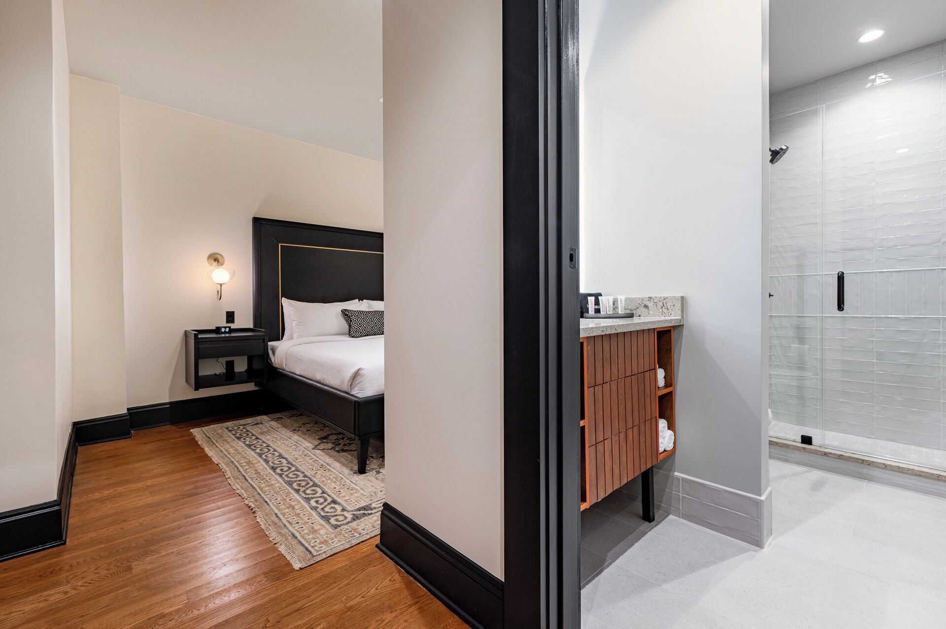 Una habitación de hotel moderna con una cama grande, una mesita de noche con lámpara, un tocador de madera y una ducha a pie.