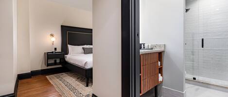 Una habitación de hotel moderna con una cama grande, una mesita de noche con lámpara, un tocador de madera y una ducha a pie.