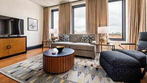 A modern hotel room with a sofa, two armchairs, a TV, and a large window with curtains.