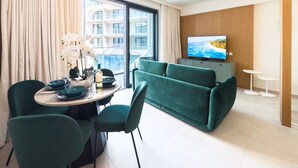 Habitación de hotel moderna con zona de comedor, sofá verde, televisor y vista panorámica de la ciudad.