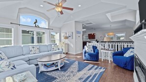 A spacious living room with a ceiling fan, a large window, a fireplace, and a kitchen area in the background. - CB49 - Cruisin' The Dunes, Gated Corolla Townhome w/ Hot Tub & Resort Pool (Corolla)