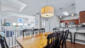 A spacious dining area with a wooden table and black chairs, a kitchen with modern appliances, and a large window providing natural light. - CB49 - Cruisin' The Dunes, Gated Corolla Townhome w/ Hot Tub & Resort Pool (Corolla)