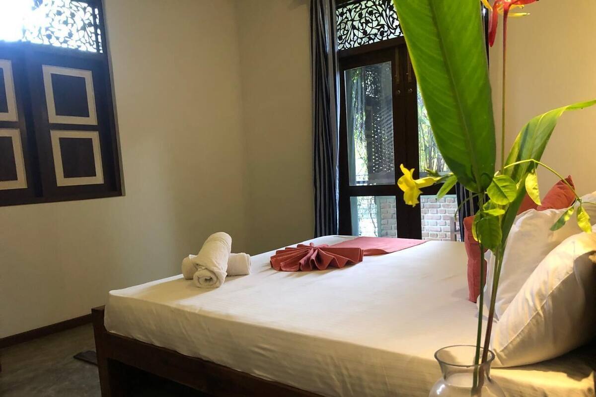A neatly made bed with a white sheet, a rolled white towel, and a pink towel. There is a clear glass vase with yellow flowers on the bedside table.