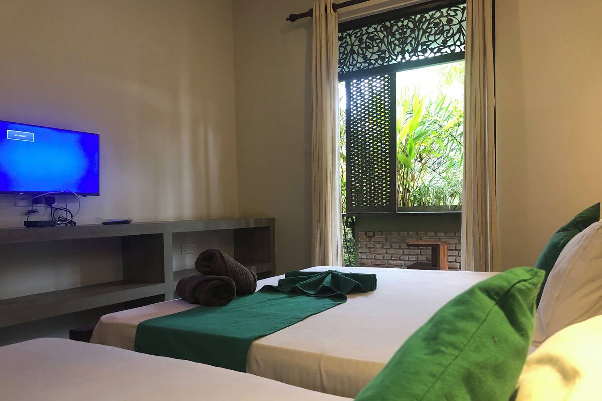 A hotel room with two beds, a TV on the wall, and a window with a view of greenery.