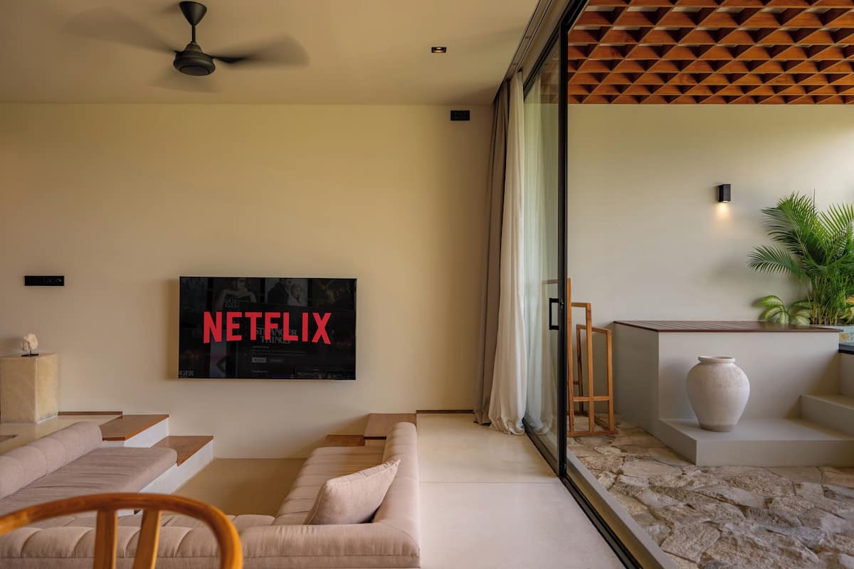 Ein modernes Wohnzimmer mit einem großen Flachbildfernseher, der 'NETFLIX' zeigt, einem beigefarbenen Sofa, einem Holzstuhl, einem Steinfußboden und einer großen Vase auf einer Plattform.