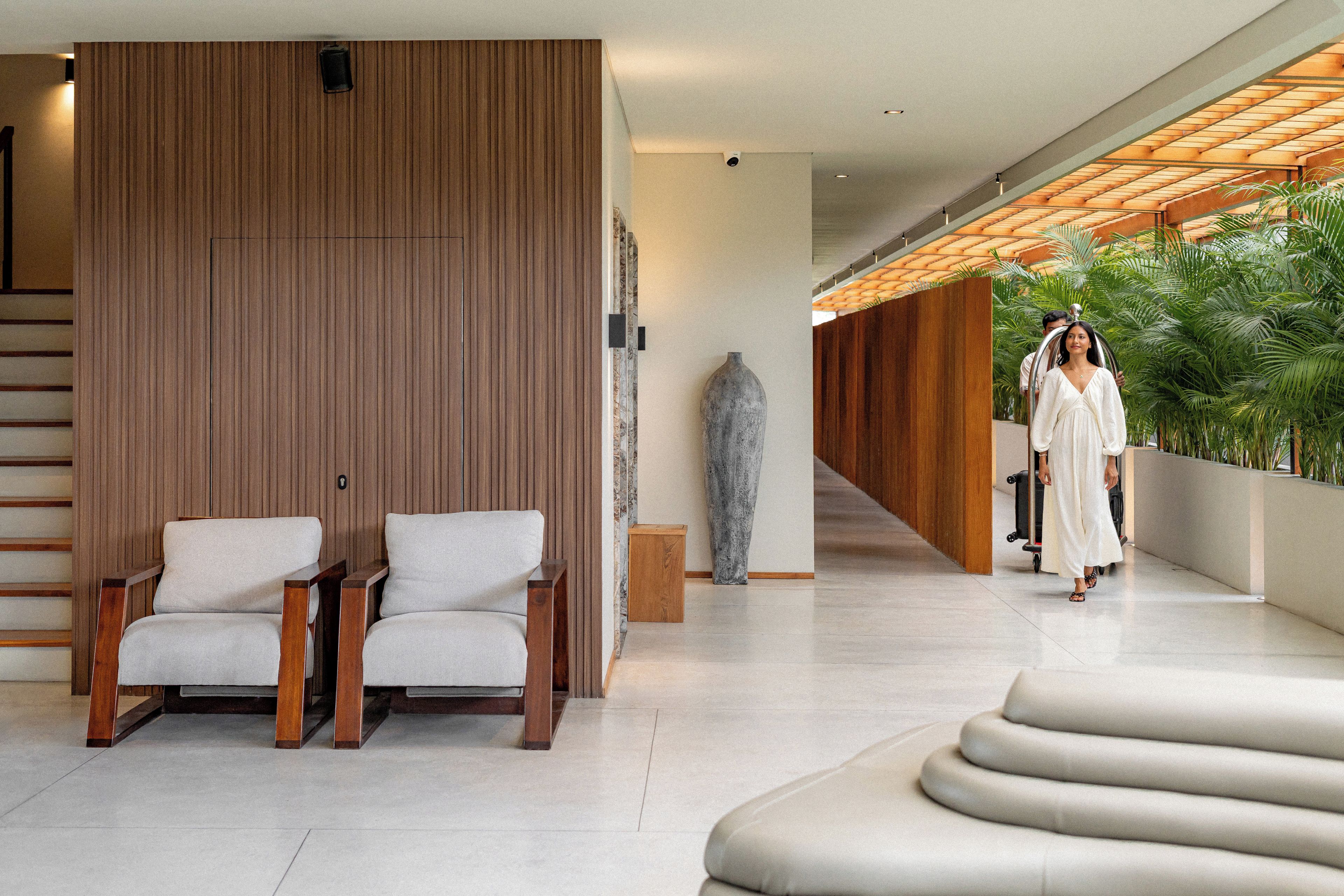 A modern interior with a wooden panel wall, two white armchairs, a large vase, and a staircase leading to a balcony with greenery.