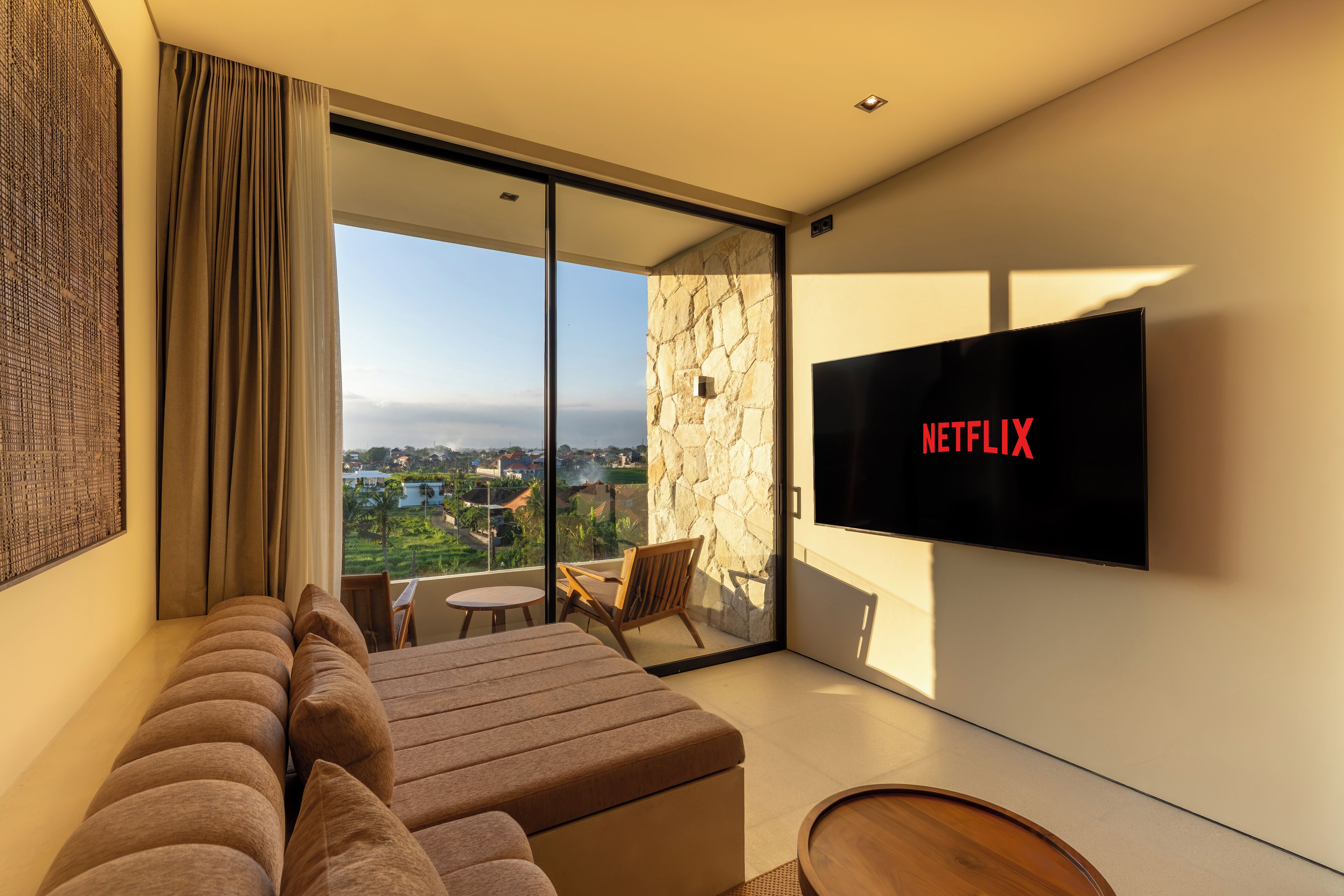 A modern hotel room with a large bed, a flat-screen TV, a balcony with a view, and a stone accent wall.