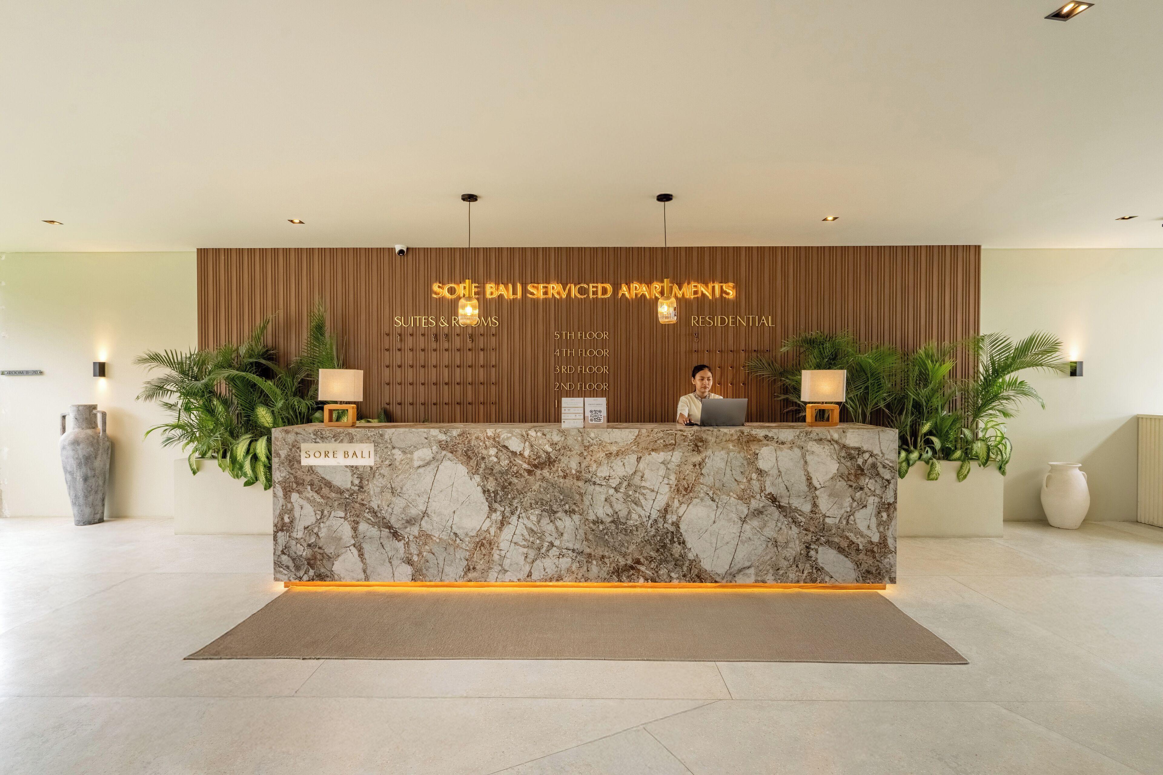 A reception desk with a marble top, two lamps, and a sign indicating 'Sole Bali Serviced Apartments' with room types listed above.
