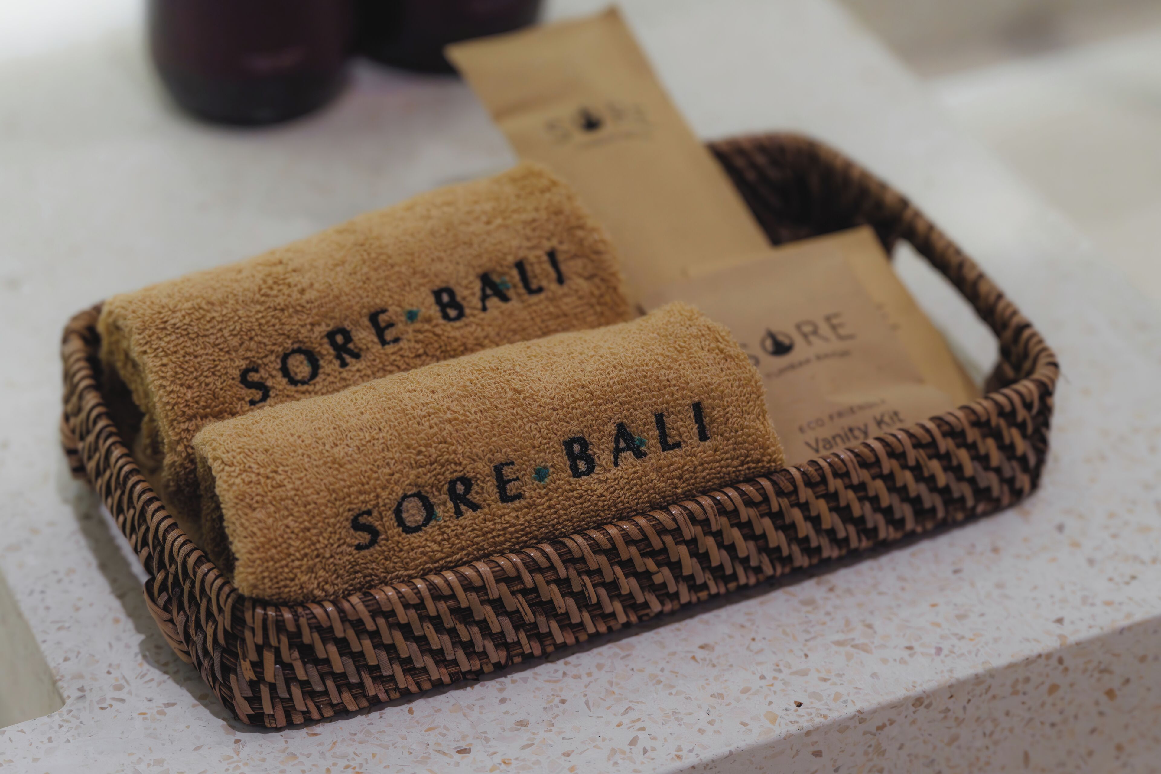 Una cesta tejida con dos toallas que dicen 'SORE-BALI' y una pequeña bolsa de papel marrón con marca similar.