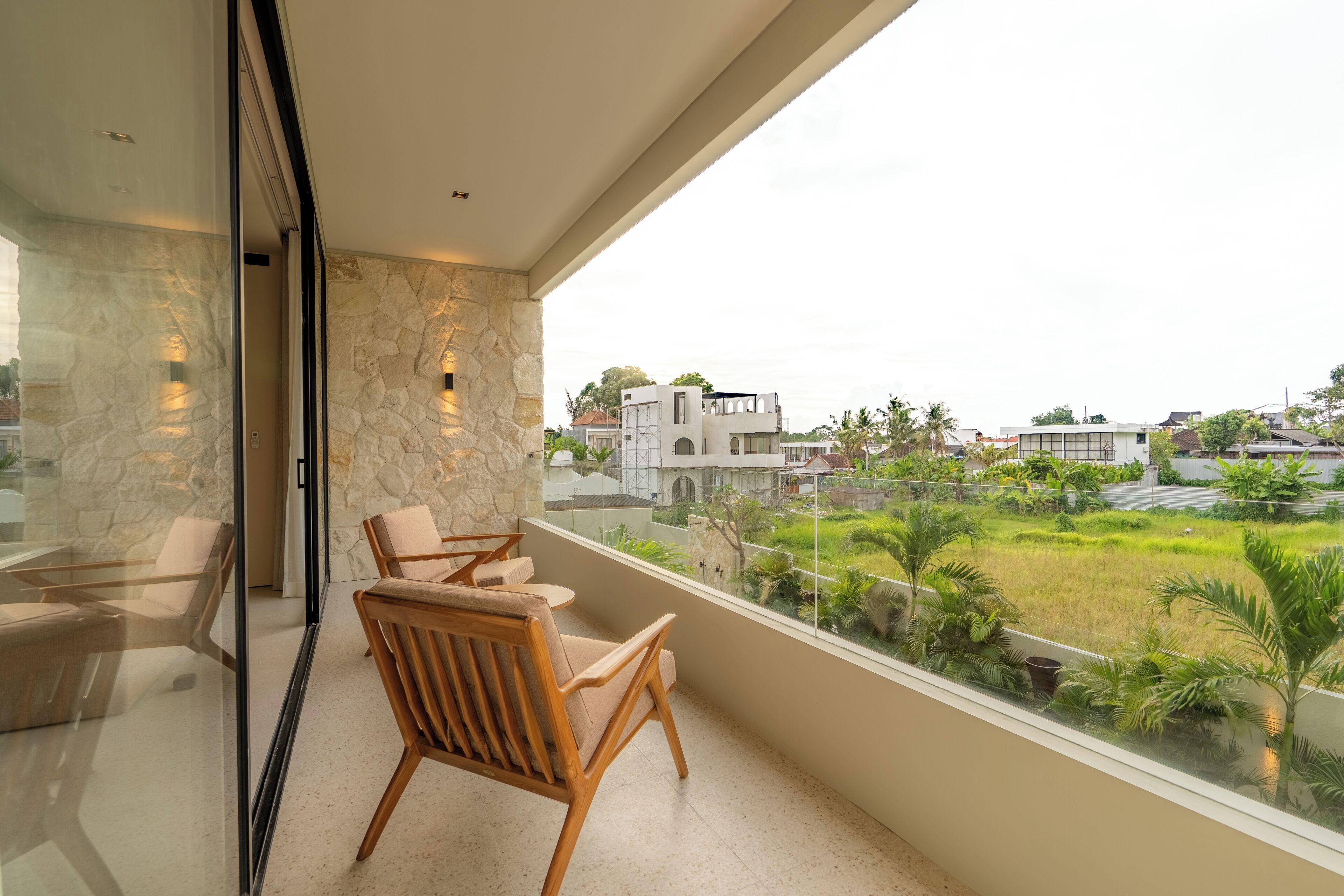 Un balcone con parete in pietra, sedie di legno e vista su una zona residenziale con case e vegetazione.