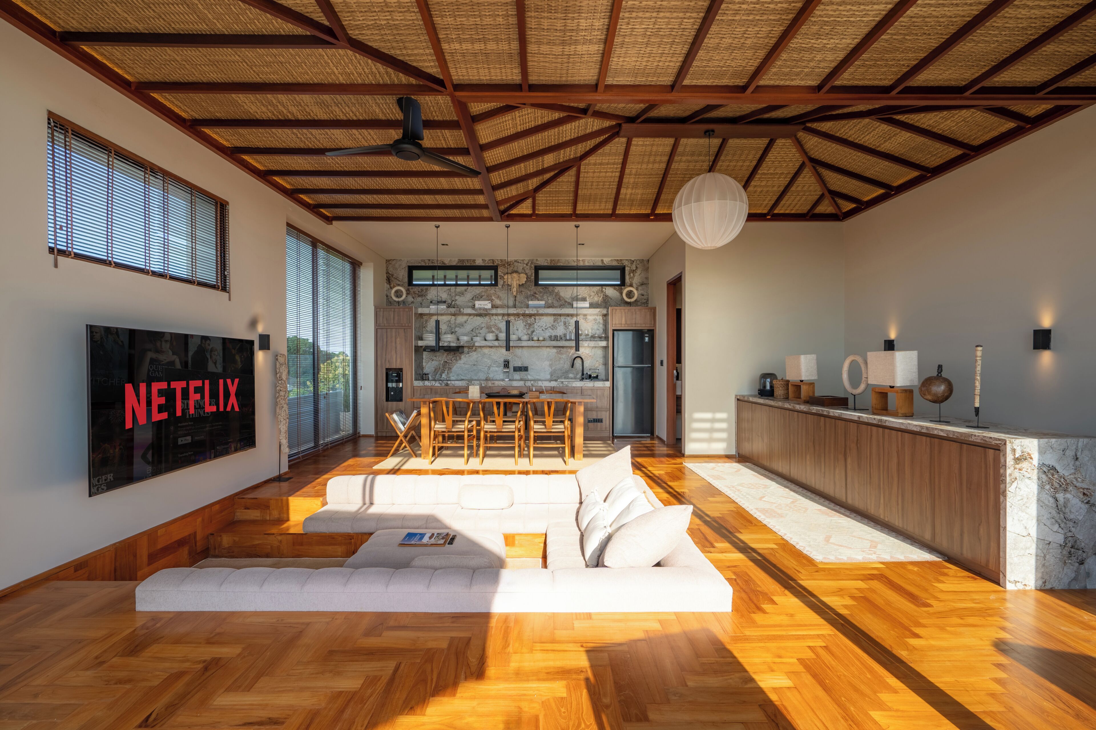 A modern living room with a large TV, wooden flooring, a sofa, and a kitchen in the background.