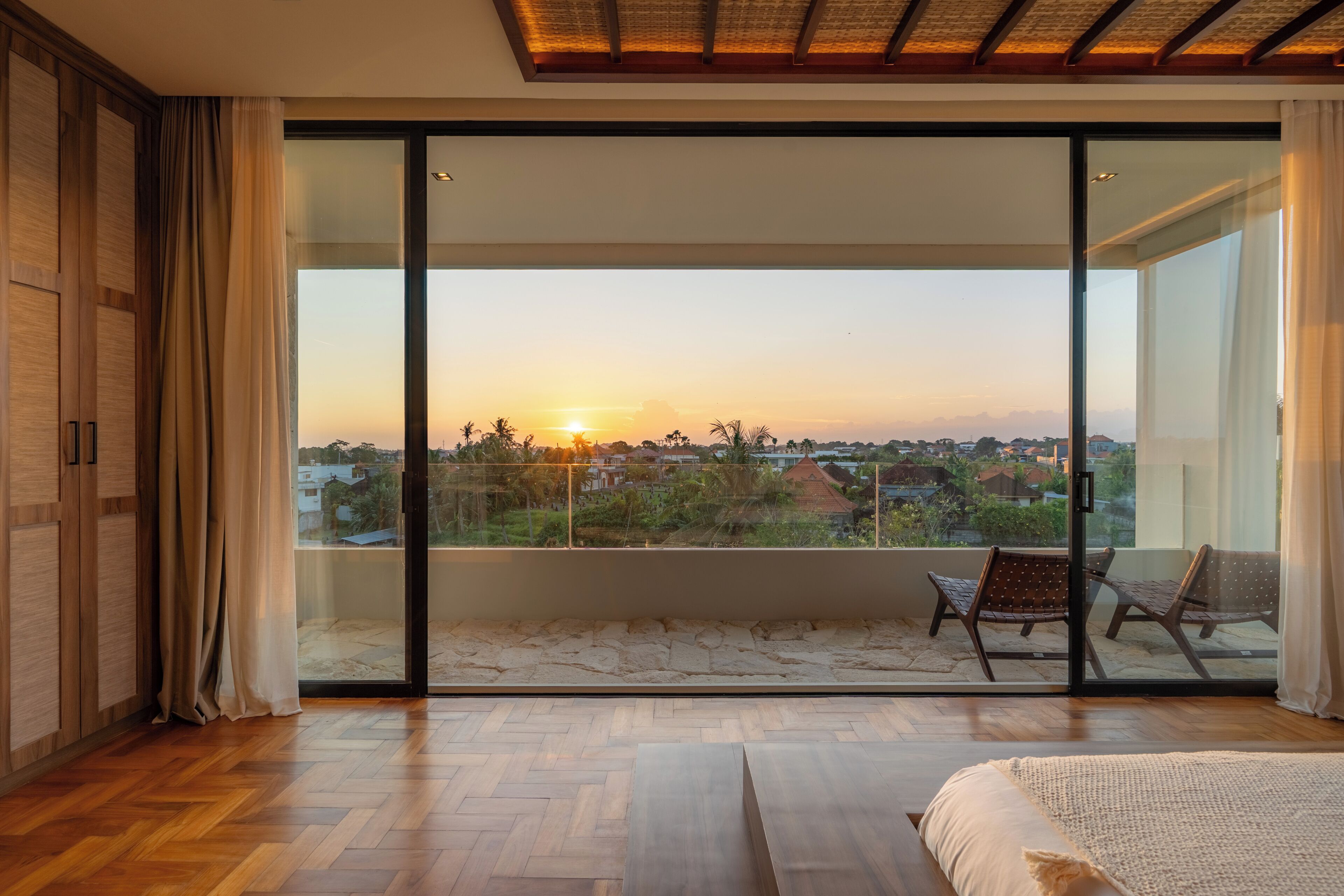 A bedroom with a large sliding glass door leading to a balcony with a view of the sunset and surrounding landscape.