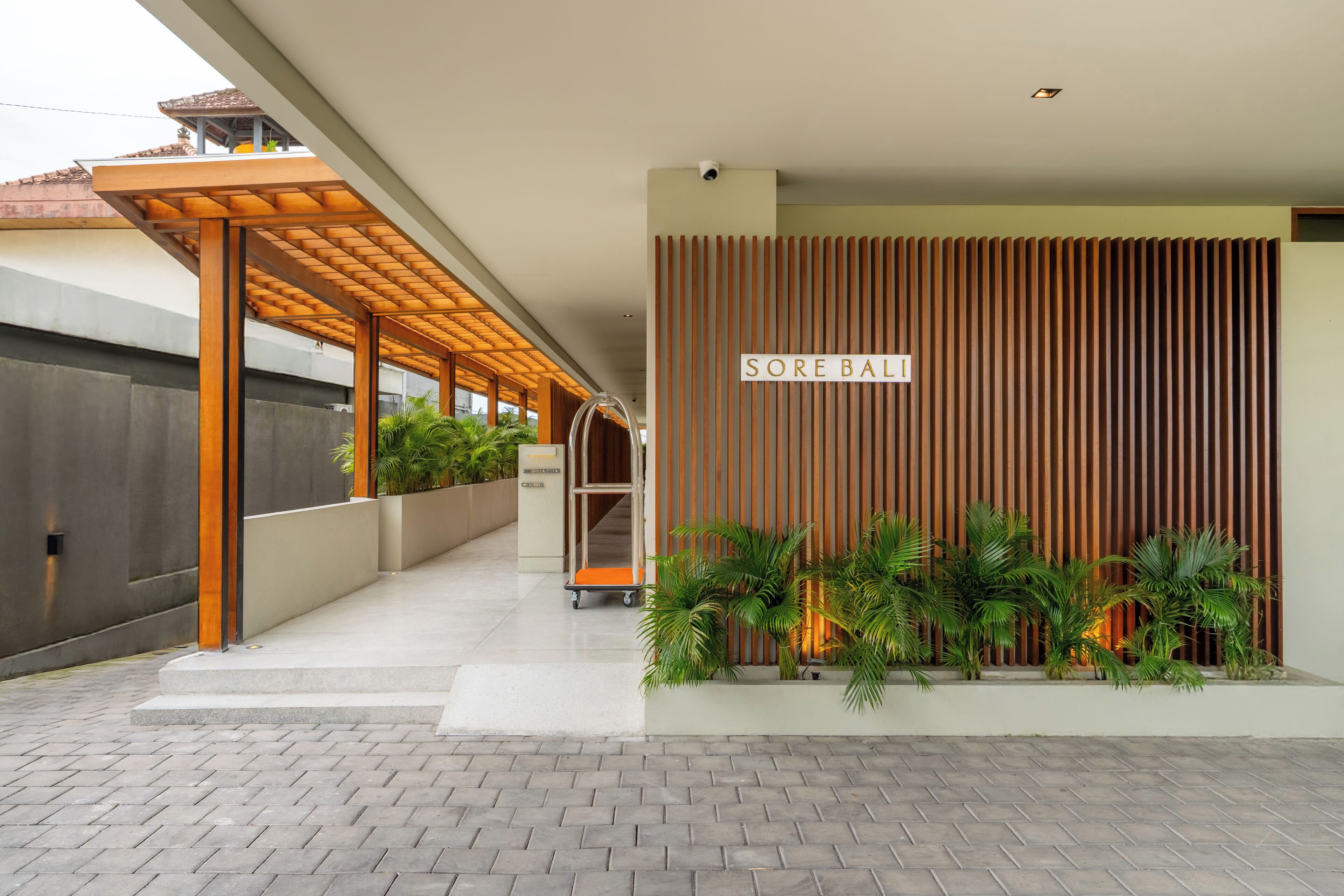 Ein moderner Hauseingang mit einer Holzpergola, einem Schild mit der Aufschrift "SORE BALI" und einem Pflanzgefäß mit Grünpflanzen.