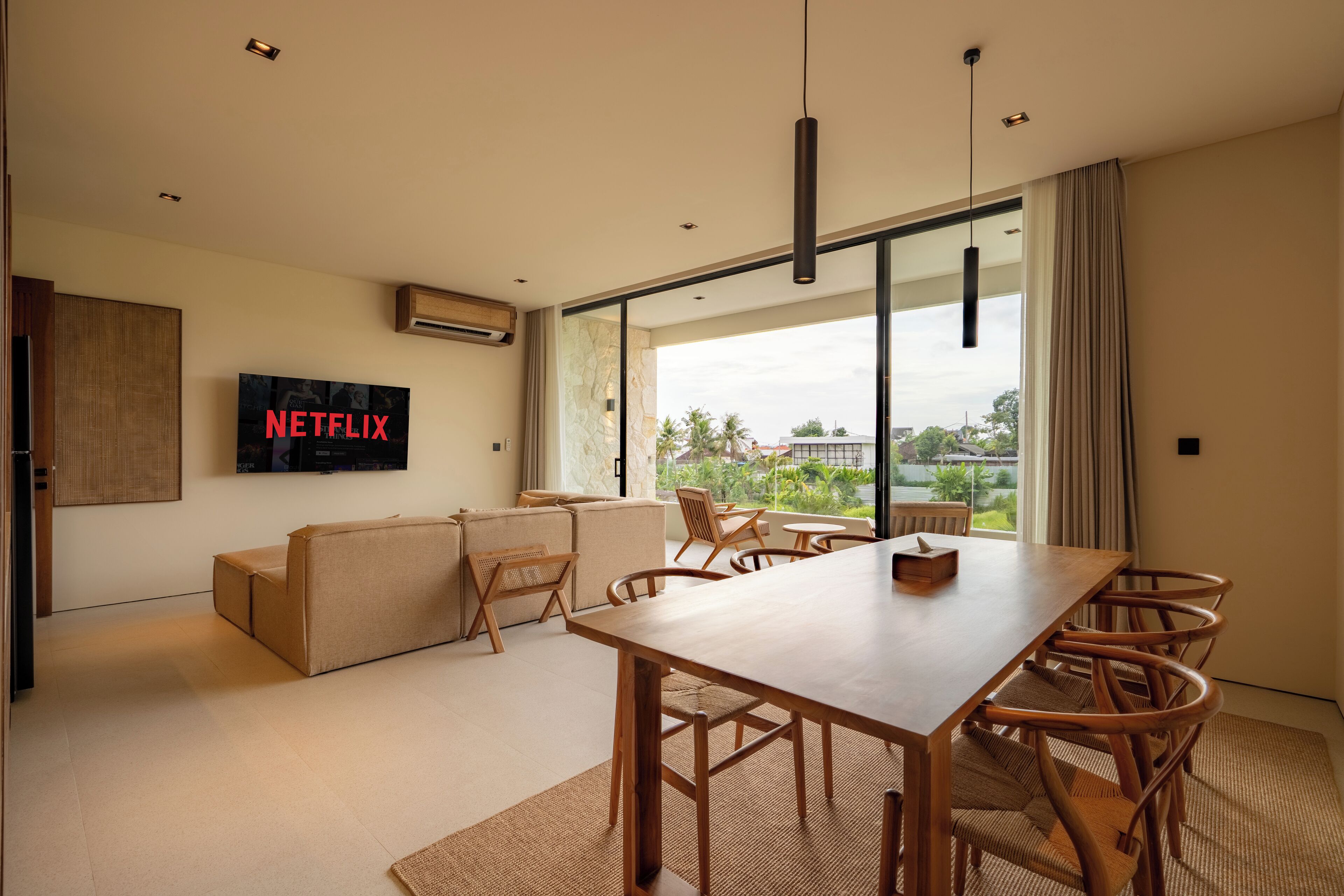 Ein modernes Wohnzimmer mit einem großen Fernseher, der 'NETFLIX' zeigt, einem Holzesstisch mit Stühlen, einem Sofa und Blick ins Freie durch große Fenster.