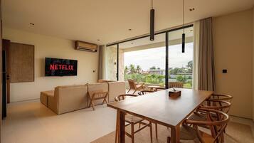 Ein modernes Wohnzimmer mit einem großen Fernseher, der 'NETFLIX' zeigt, einem Holzesstisch mit Stühlen, einem Sofa und Blick ins Freie durch große Fenster.