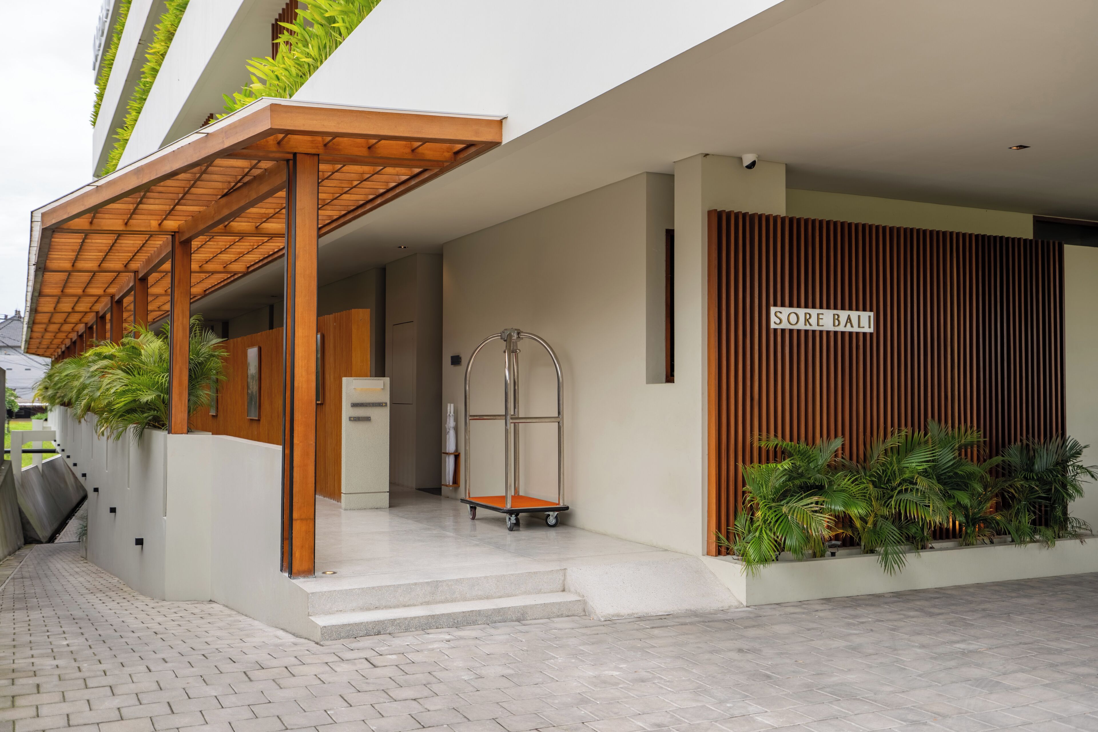Uma entrada de prédio moderna com uma pérgula de madeira, uma placa com a inscrição "SORE BALI" e um carrinho.
