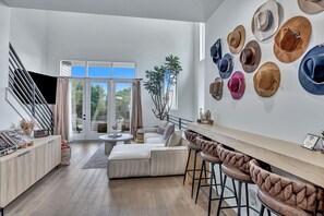 Una sala moderna con un sofá, un comedor con taburetes de bar, una escalera y una pared decorada con sombreros.