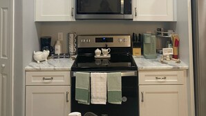 Una cocina con armarios blancos, un microondas negro y una estufa. Encima de la estufa hay cuadros con imágenes de utensilios de cocina.