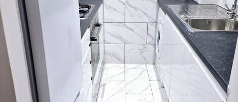 A compact kitchen with white marble tiles, a built-in gas stove, a stainless steel sink, and a white refrigerator.