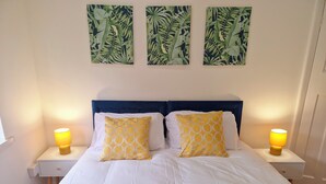 Un lit avec une literie blanche et deux oreillers jaunes, de chaque côté duquel se trouvent des tables de chevet avec des lampes, dans une chambre dotée d’une tête de lit bleue et de trois œuvres d’art murales représentant des motifs de feuilles, placées au-dessus du lit.