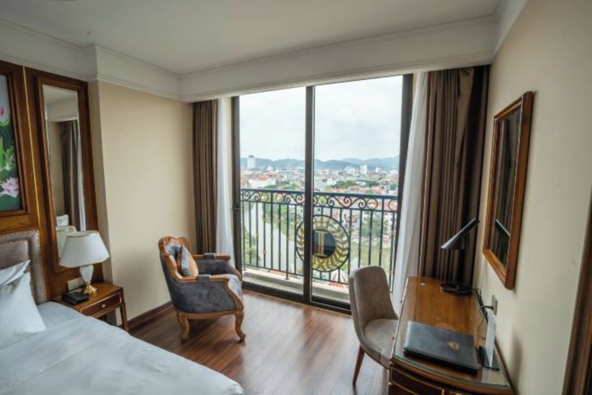 Ein Hotelzimmer mit einem großen Bett, zwei Sesseln, einem Schreibtisch mit einem Laptop und einem Balkon mit Blick auf die Stadt.