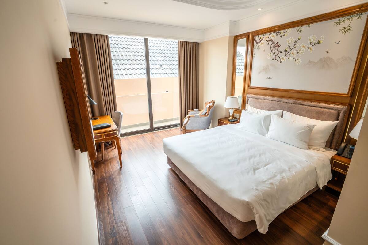 A hotel room with a large bed, a desk with a chair, a small table with a lamp, and a view of the outside through a sliding door.