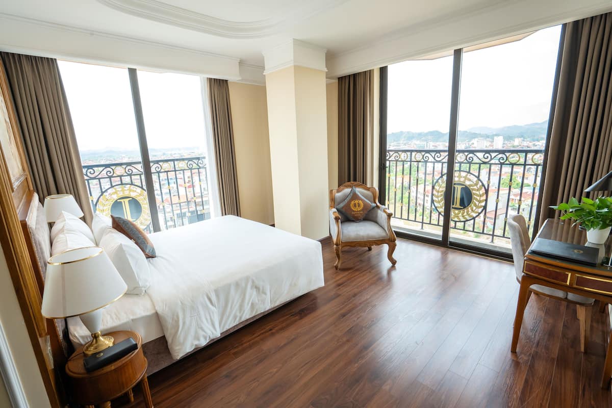 A hotel room with a large bed, a chair, a desk, and a view of the city through large windows.