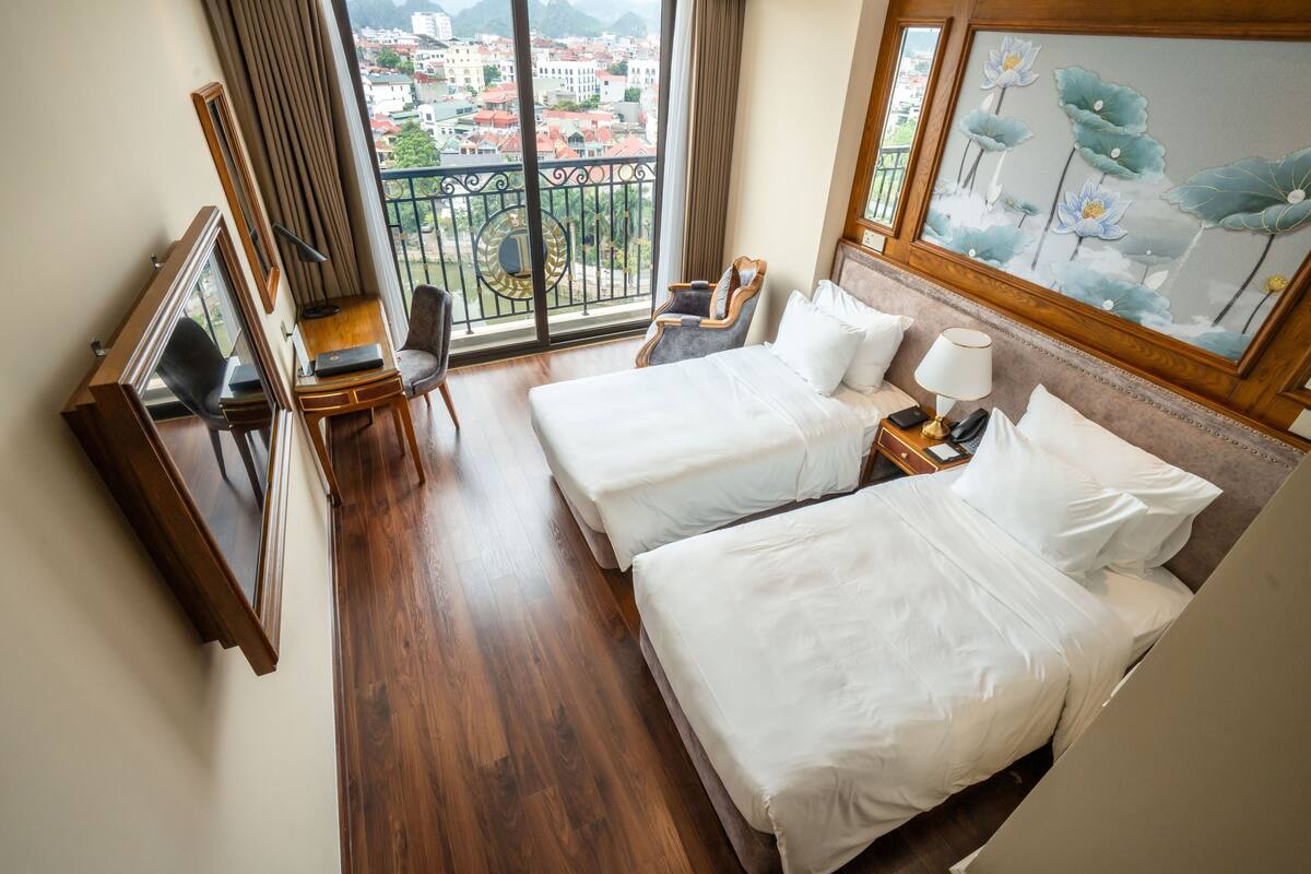 A hotel room with two beds, a desk with a chair, a TV, and a large window with a city view.