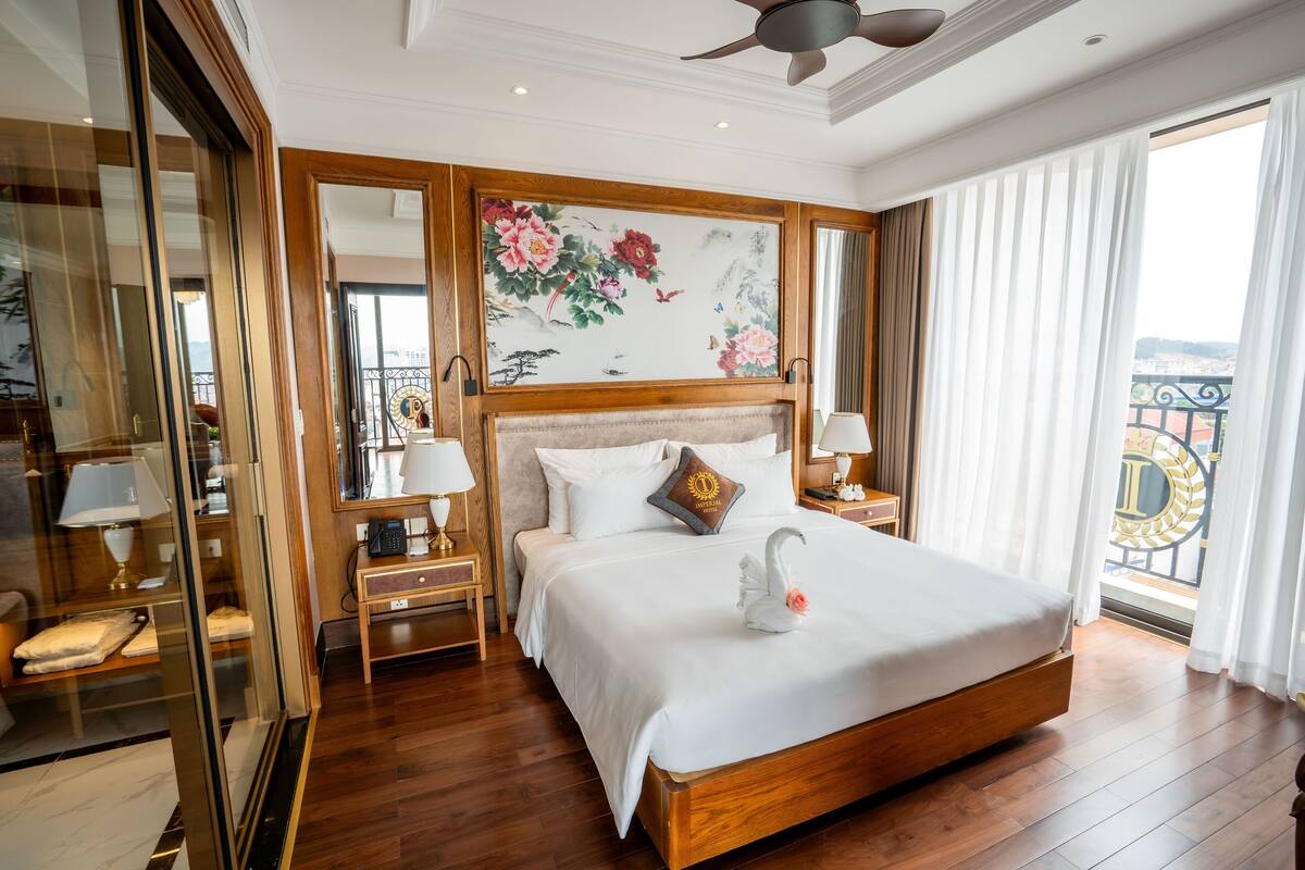 A hotel room with a large bed, wooden headboard, a painting on the wall, and a balcony with a view.