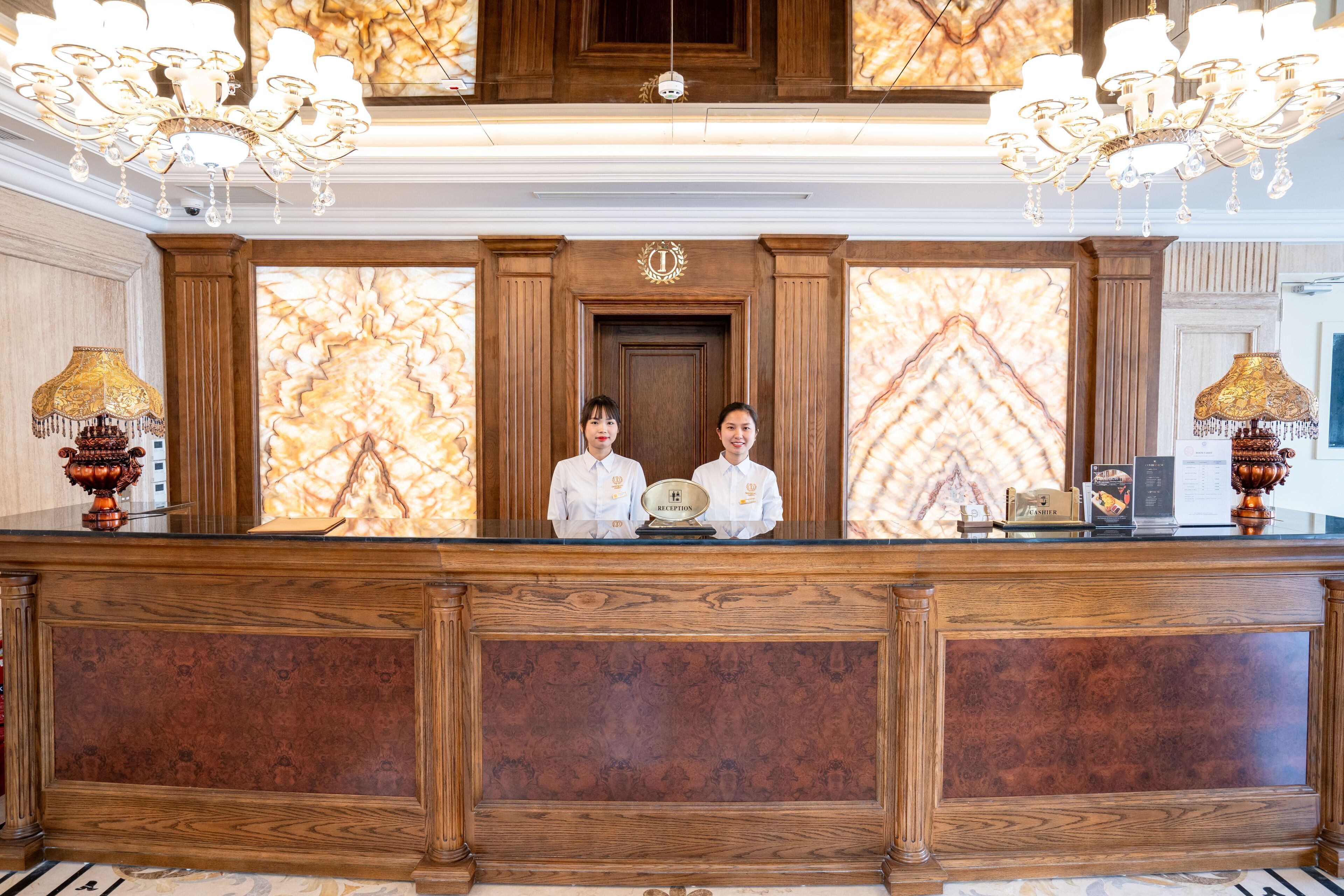A reception desk with two staff members, a wooden panel design, and a chandelier.
