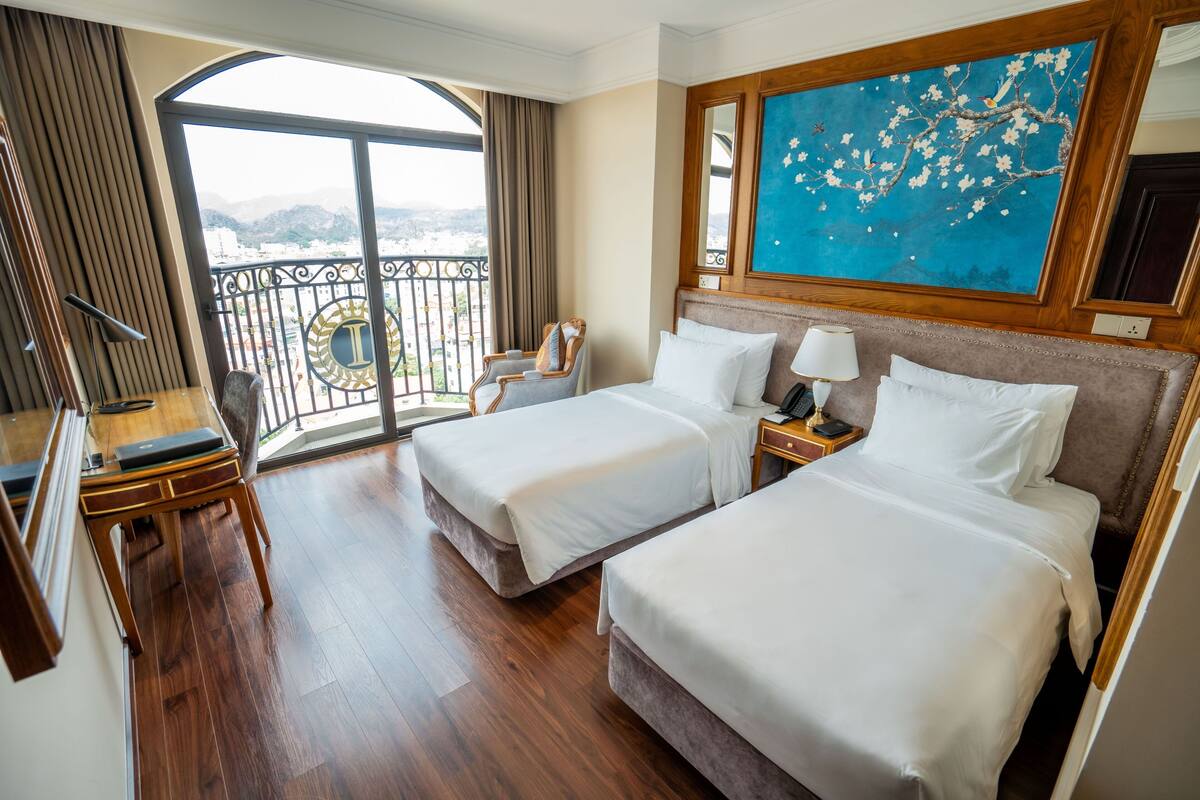 A hotel room with two beds, a desk, a chair, a large window with a view of mountains, and a painting on the wall.