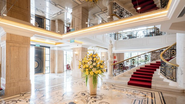 Un amplio vestíbulo con piso de mármol, una escalera con alfombra roja y un gran jarrón con flores amarillas.