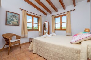 A bedroom with a large bed, two windows with curtains, a wicker chair, a small table, and a painting on the wall. - REYNES - Rustic villa with private pool and free Wi-Fi (Pollença)