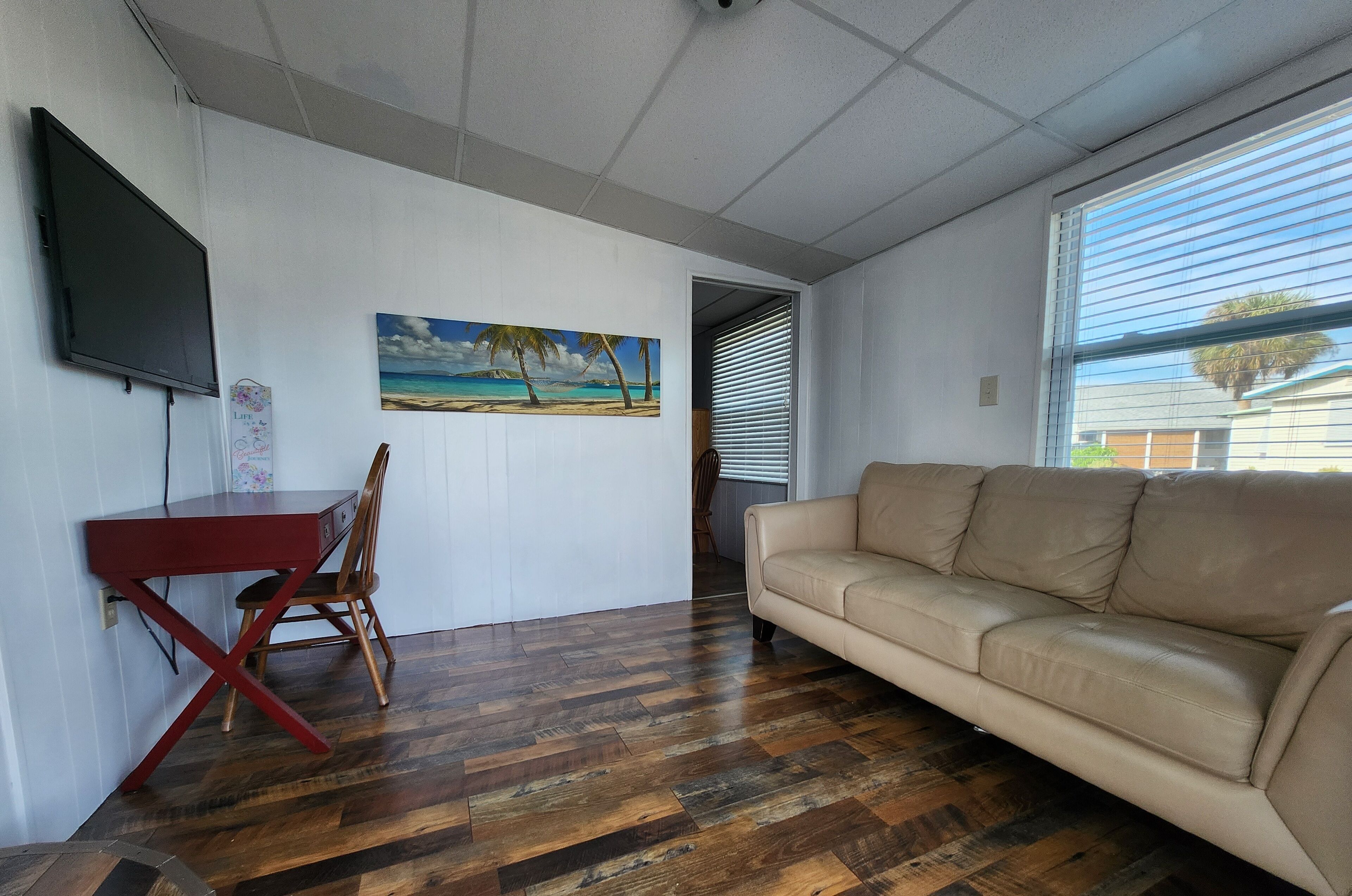 Uma sala de estar com piso de madeira, um sofá bege, uma mesa vermelha com uma cadeira, uma TV fixada na parede e um quadro com tema de praia.