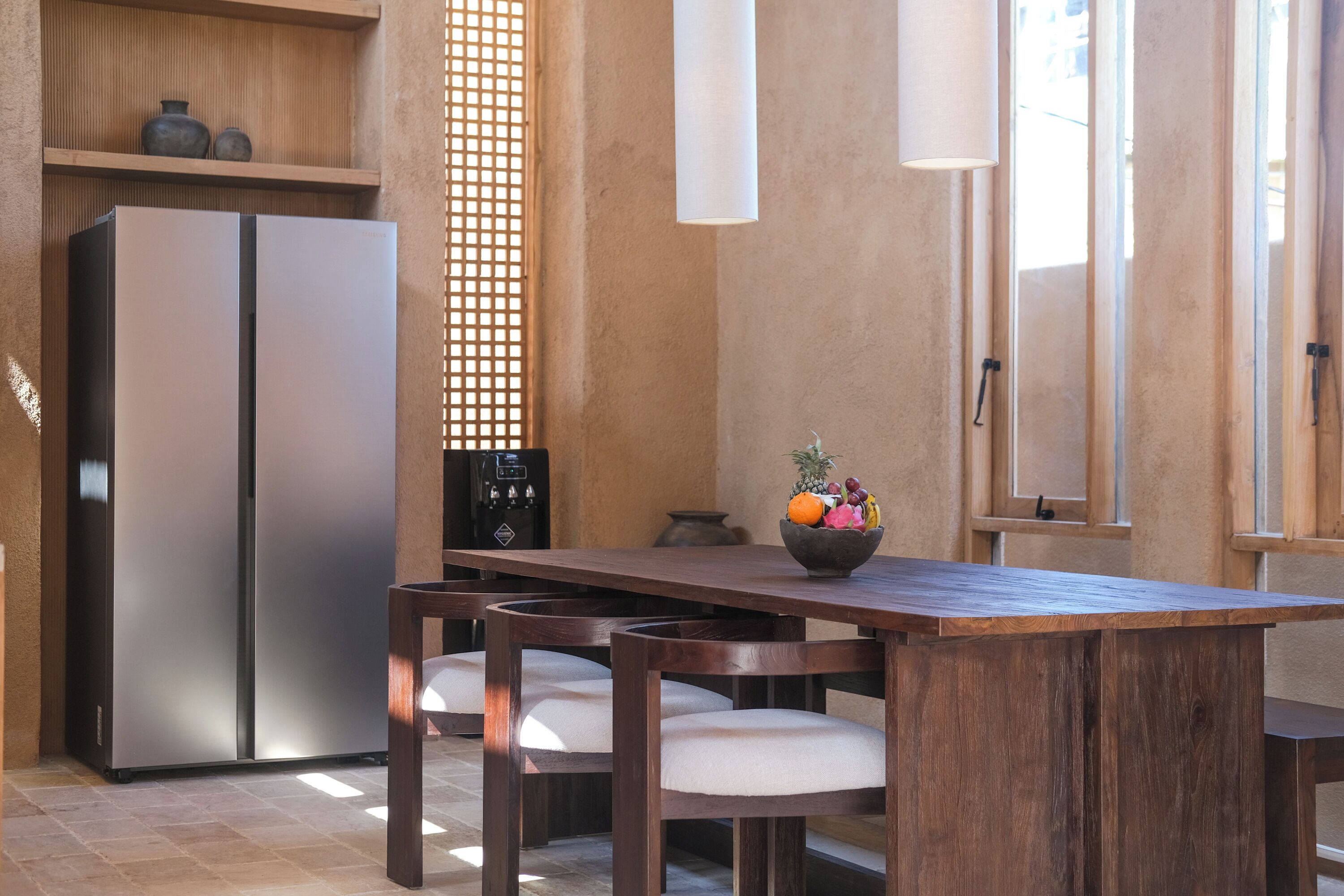 A kitchen with a wooden table, two chairs, a refrigerator, and a fruit bowl.