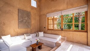 Una habitación con pared beige texturizada, un ventanal con vista a follaje, un sofá seccional beige y una mesa de centro de madera con un florero.