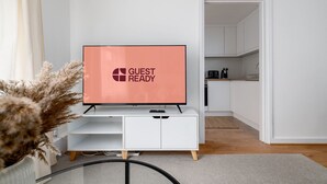 Een moderne woonkamer met een flatscreen-tv waarop “GUEST READY” staat, een witte entertainmentkast, een keuken op de achtergrond en een decoratieve plant.